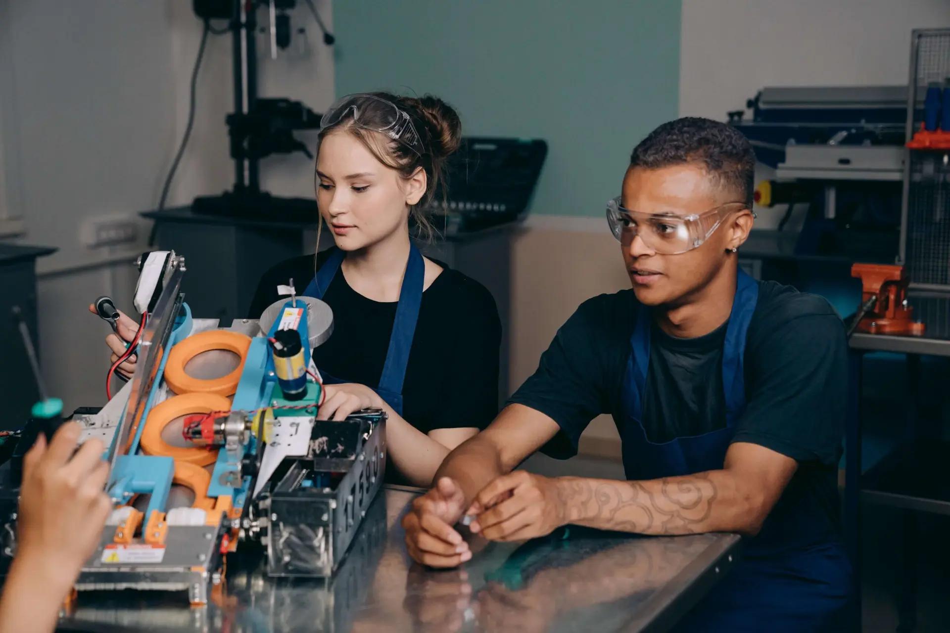 Educação corporativa para a nova era da indústria e inovação Dois jovens usando óculos de proteção montam um equipamento robótico industrial sobre uma bancada metálica em laboratório.