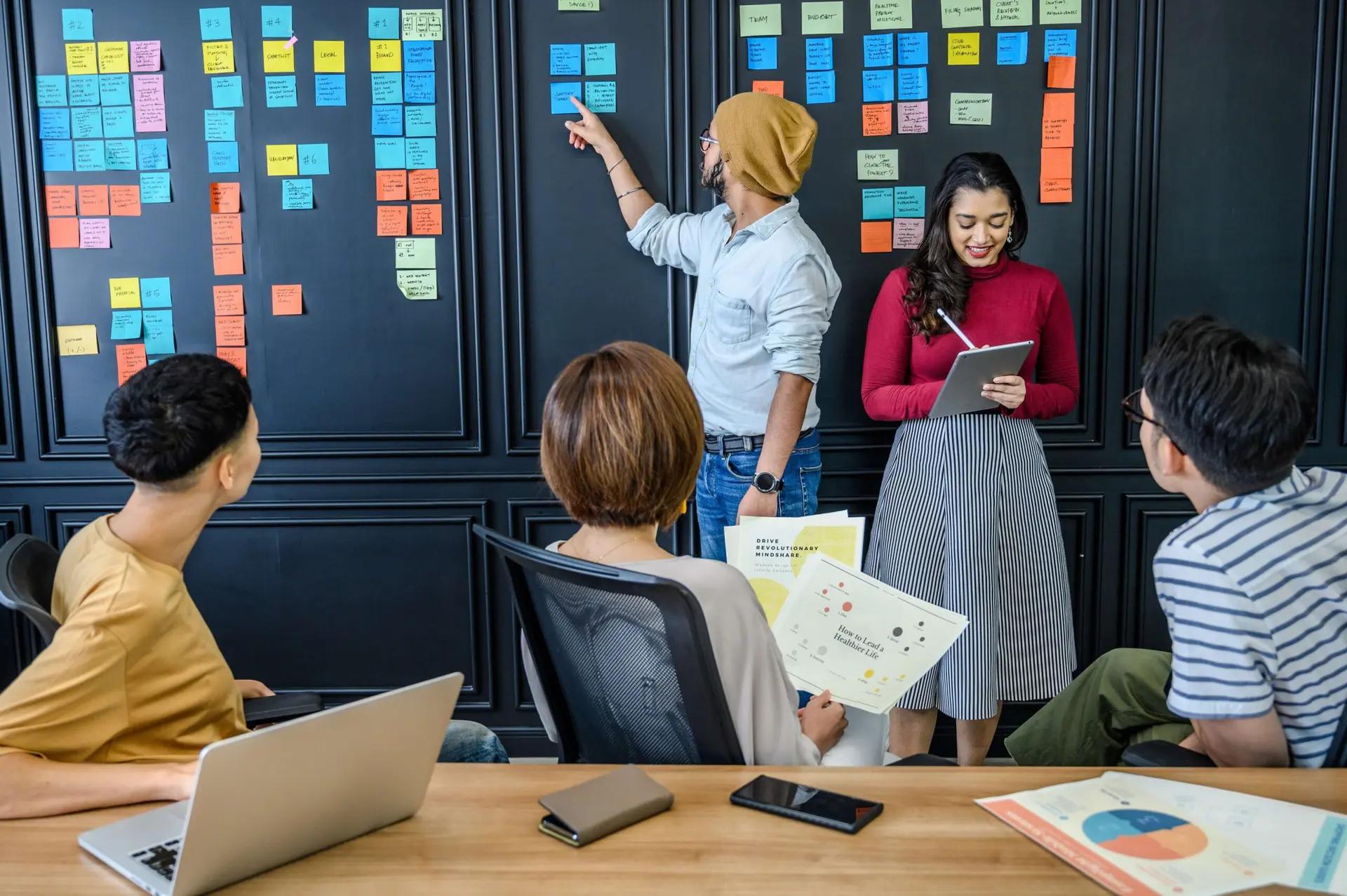 Tipos de Inovação – Conheça os Modelos e Como Aplicar em seu Negócio Cinco pessoas em reunião de planejamento em frente a quadro negro repleto de post-its coloridos, usando notebook e tablet.
