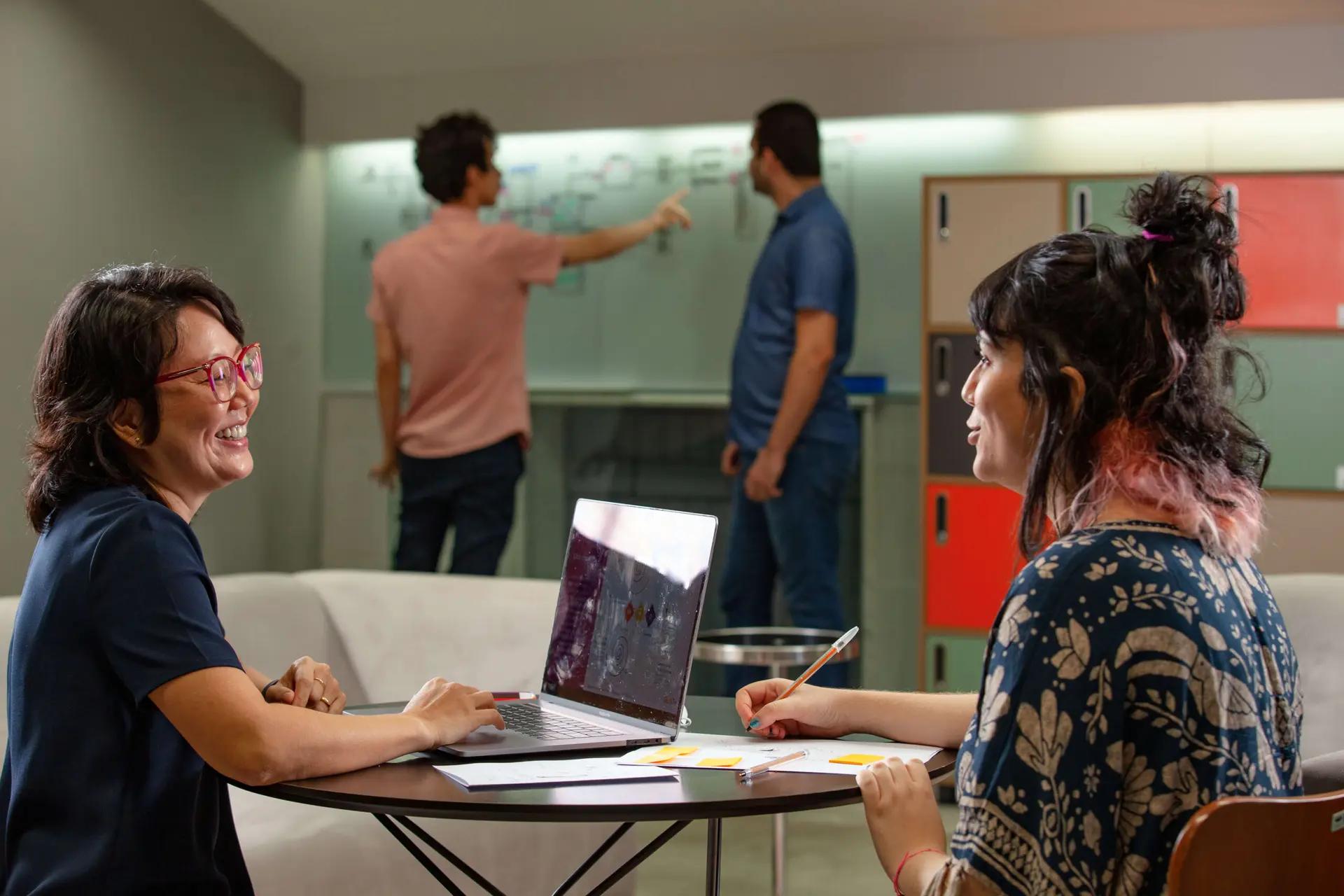 Intraempreendedores – Como desenvolver talentos dentro da sua empresa Duas mulheres conversam em uma mesa redonda com laptop e papéis; ao fundo, dois homens discutem diante de um quadro branco.