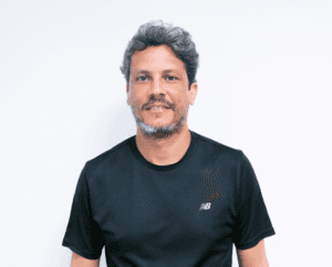 Homem com cabelo e barba grisalhos curtos, vestindo uma camisa esportiva preta, em pé contra um fundo branco.
