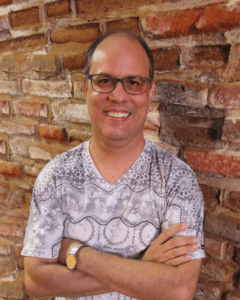 Homem sorridente, usando óculos e uma camisa estampada, em pé, encostado em uma parede de tijolos.