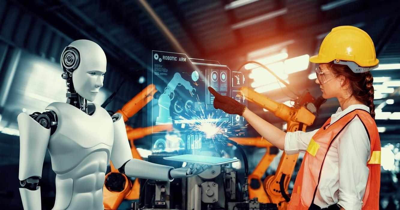 Imagem de capa Uma mulher de capacete e óculos de segurança interage com um robô humanóide e um painel de controle holográfico em uma fábrica futurista. Braços robóticos industriais são visíveis ao fundo.
