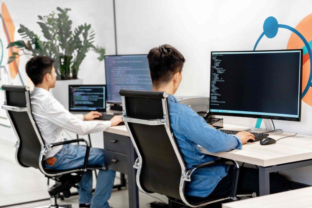 Dois programadores trabalhando em mesas com vários monitores exibindo códigos em um espaço de escritório moderno, com plantas ao fundo.