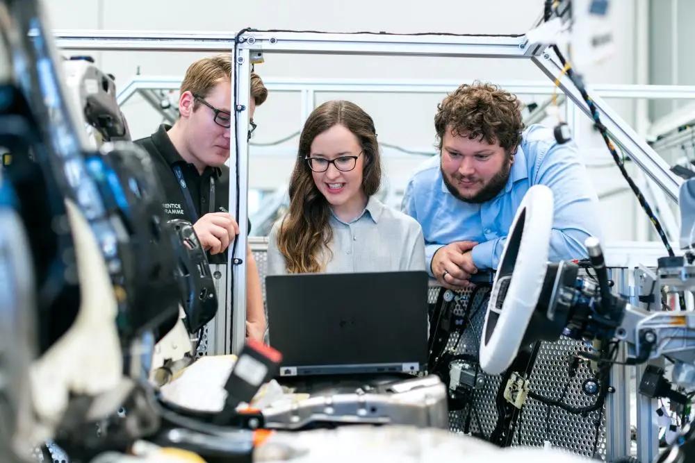 EMBRAPII – como captar recursos para projetos de inovação industrial Três engenheiros sorridentes analisam dados em um laptop dentro de um laboratório industrial cercados por cabos e máquinas.
