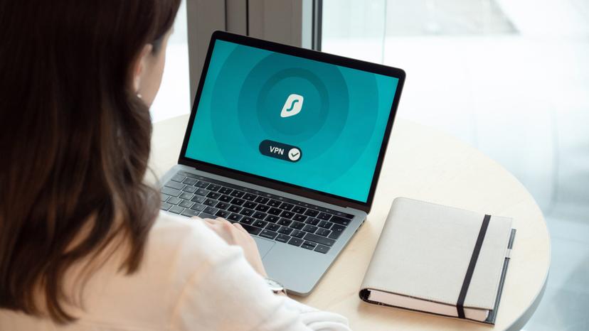 pexels-dan-nelson-3949100 Uma mulher com cabelo castanho está sentada à mesa usando um laptop. A tela mostra uma interface de VPN ativada. Um caderno está ao lado do laptop.