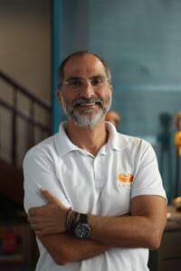 Homem sorridente, de óculos e barba, vestindo uma camisa polo branca, braços cruzados, em pé em ambiente interno com fundo desfocado.