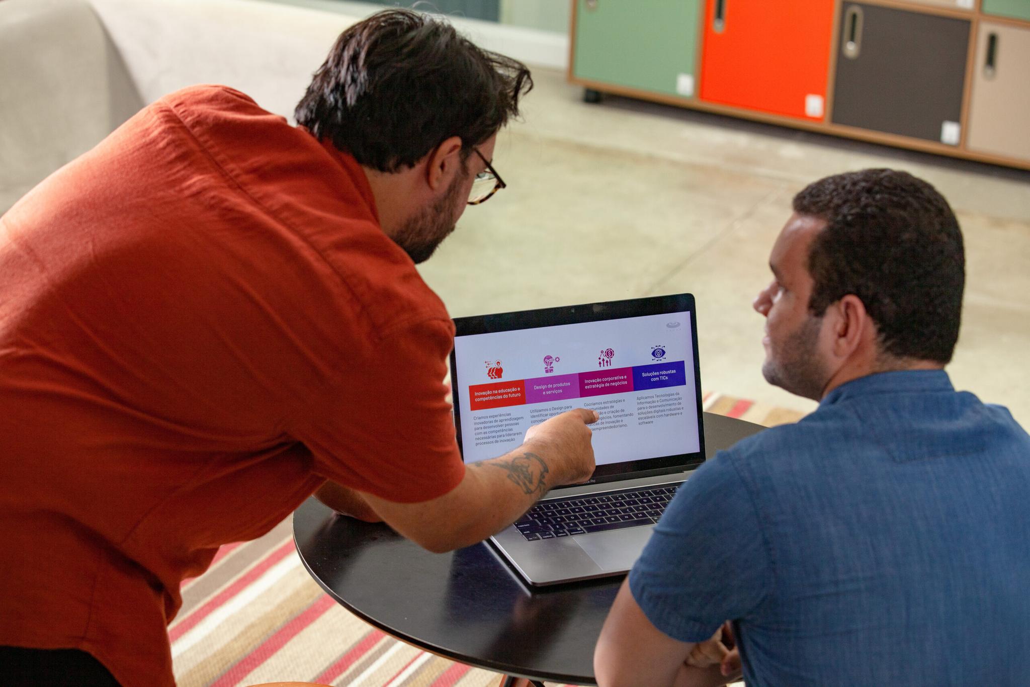 _MG_3430 Dois homens analisam juntos uma apresentação em um laptop. Um homem de camisa laranja aponta para a tela, enquanto o outro, de camisa azul, observa atentamente.