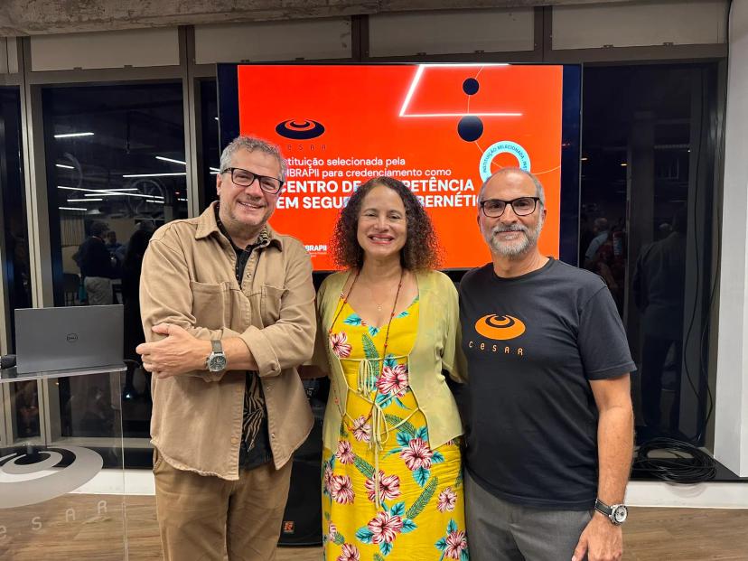 centro-ciberseguranca Três pessoas posam sorrindo para uma foto em frente a uma tela com um anúncio de credenciamento.