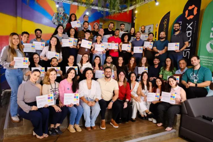 Formação em agilidade para servidores públicos Grupo de aproximadamente 40 servidores sorridentes exibe certificados coloridos em um ambiente moderno e vibrante