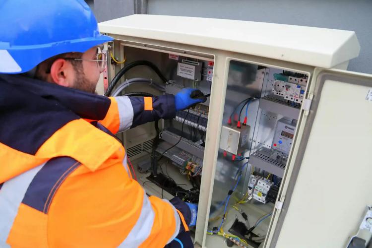 Modernização de Sistemas Legados no Setor de Energia Técnico com capacete azul e colete laranja a ajustar cabos num armário de distribuição elétrica aberto com diversos circuitos.