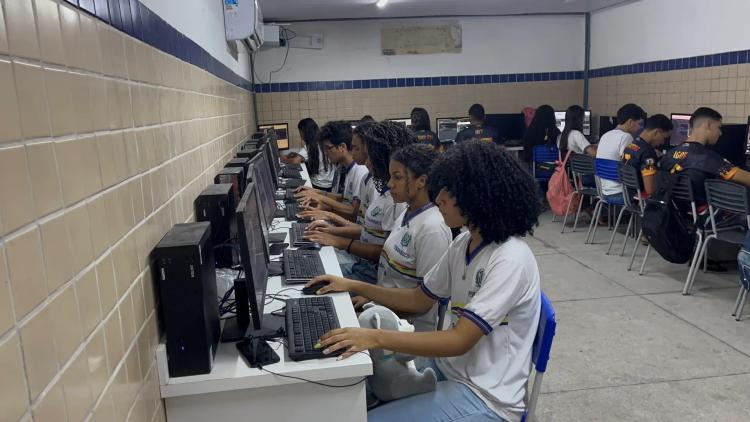Impacto Social na Educação e Conexão de Estudantes ao Mercado Tech Grupo de jovens sentados em bancadas usando computadores desktop em um laboratório de informática escolar com paredes de azulejo.